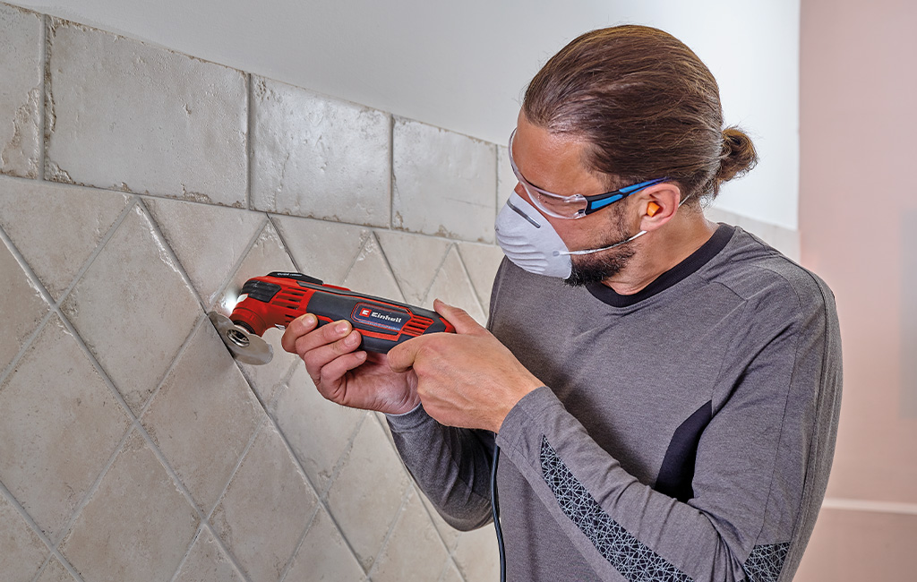 A man wearing protective gear uses an Einhell multitool to remove grout from tiled walls.