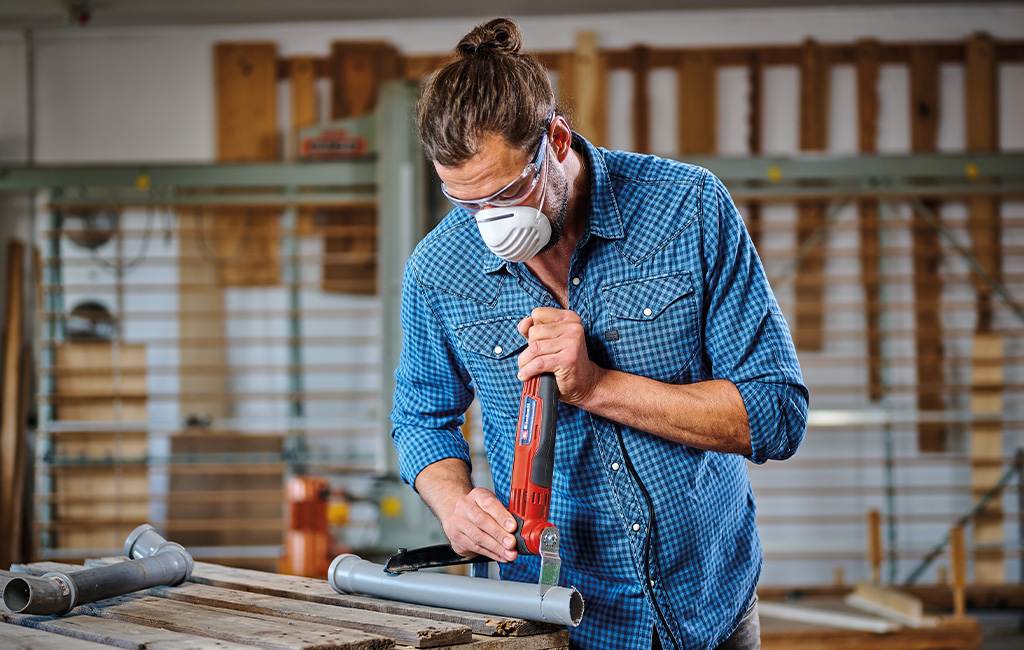 Muškarac reže plastičnu cijev pomoću Einhell multitoola te pritom nosi zaštitne naočale i masku.