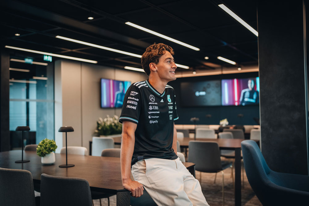 George Russell, vozač Mercedes-AMG PETRONAS Formula One tima, opušteno sjedi na stolu u modernom i elegantnom prostoru za goste ili uredu tima. Smiješi se i nosi službenu majicu tima s vidljivim logotipima sponzora kao što su Einhell, IWC i CrowdStrike. Na ekranima u pozadini prikazana je njegova slika.