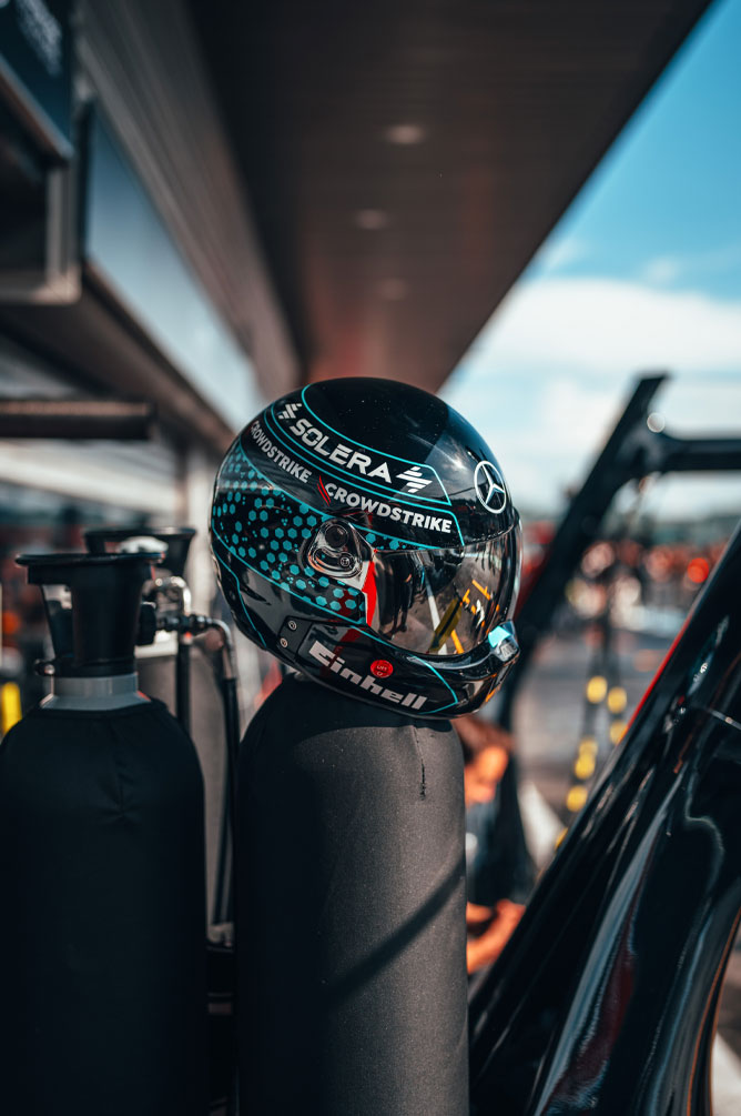 Kaciga Mercedes-AMG PETRONAS Formula One tima s jasno vidljivim logotipima sponzora, uključujući Einhell, CrowdStrike i Solera, postavljena je na bocu s plinom u boksu. Odsjaj na viziru prikazuje okruženje garaže.
