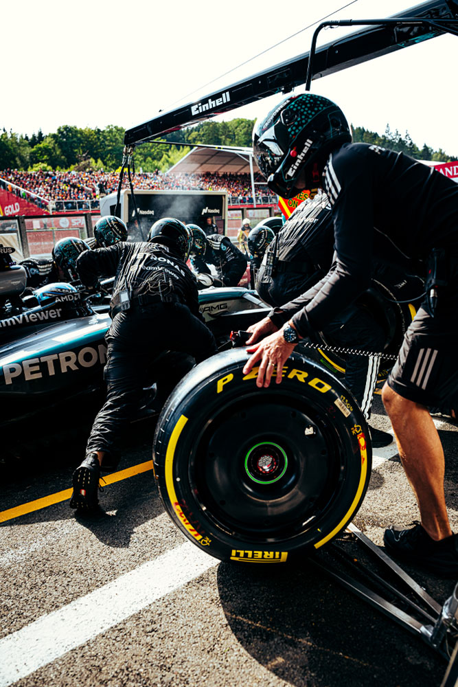 Mehaničari Mercedes-AMG PETRONAS Formula One tima izvode zaustavljanje u boksu. Jedan član tima priprema Pirelli gumu dok drugi rade na automobilu. Einhell logo vidljiv je na opremi iznad automobila.