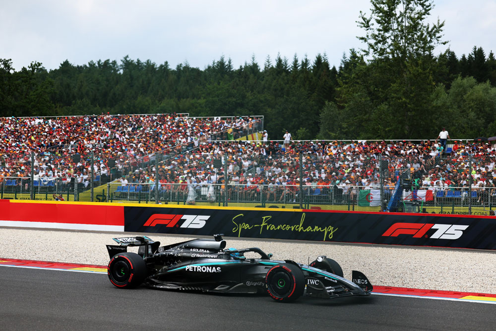 Automobil Mercedes-AMG PETRONAS Formula One tima vozi se stazom Spa-Francorchamps. U pozadini su pune tribine s navijačima koji mašu međunarodnim zastavama. Logo Spa-Francorchampsa vidljiv je na barijeri.