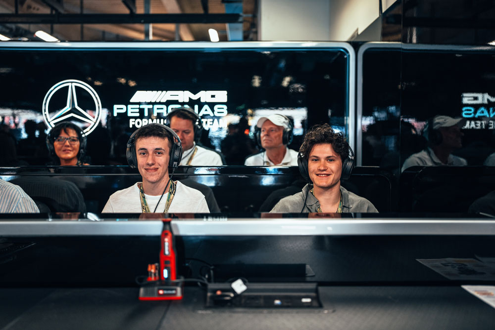 Dva muškarca sjede u garaži Mercedes-AMG PETRONAS Formula One tima, nose slušalice i uživaju u iskustvu iza kulisa. U prvom planu vidi se crveni Einhell uređaj.