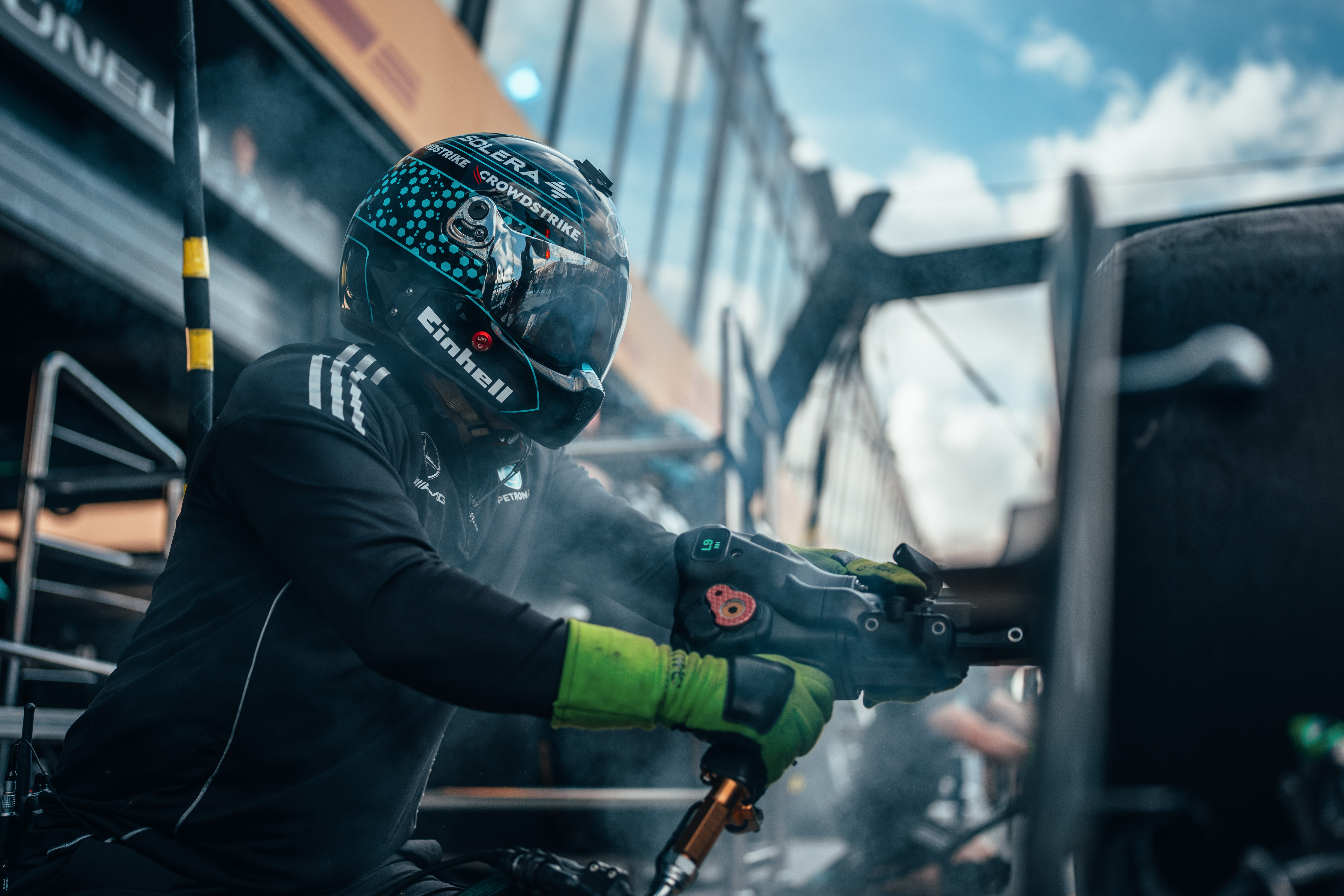 Član boks tima Formule 1, s kacigom i rukavicama, koristi pneumatski pištolj za kotače tijekom zaustavljanja, okružen dimom i opremom.