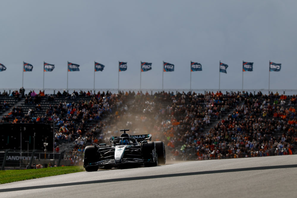 Automobil Mercedes-AMG PETRONAS Formula One tima prolazi pokraj punih tribina tijekom utrke. Navijači mašu zastavama u pozadini, stvarajući živahnu atmosferu.