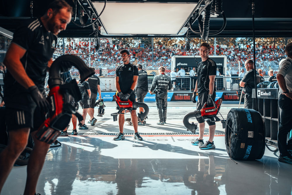 Mehaničari Mercedes-AMG PETRONAS Formula One tima pripremaju se u garaži s bežičnim puhačima Einhell prije zaustavljanja u boksu. U pozadini su tribine pune navijača.
