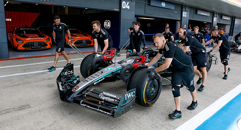 Mehrere Teammitglieder schieben einen Mercedes-AMG PETRONAS Formel-1-Rennwagen aus der Boxengasse, im Hintergrund zwei orangefarbene Mercedes-AMG GT-Fahrzeuge.