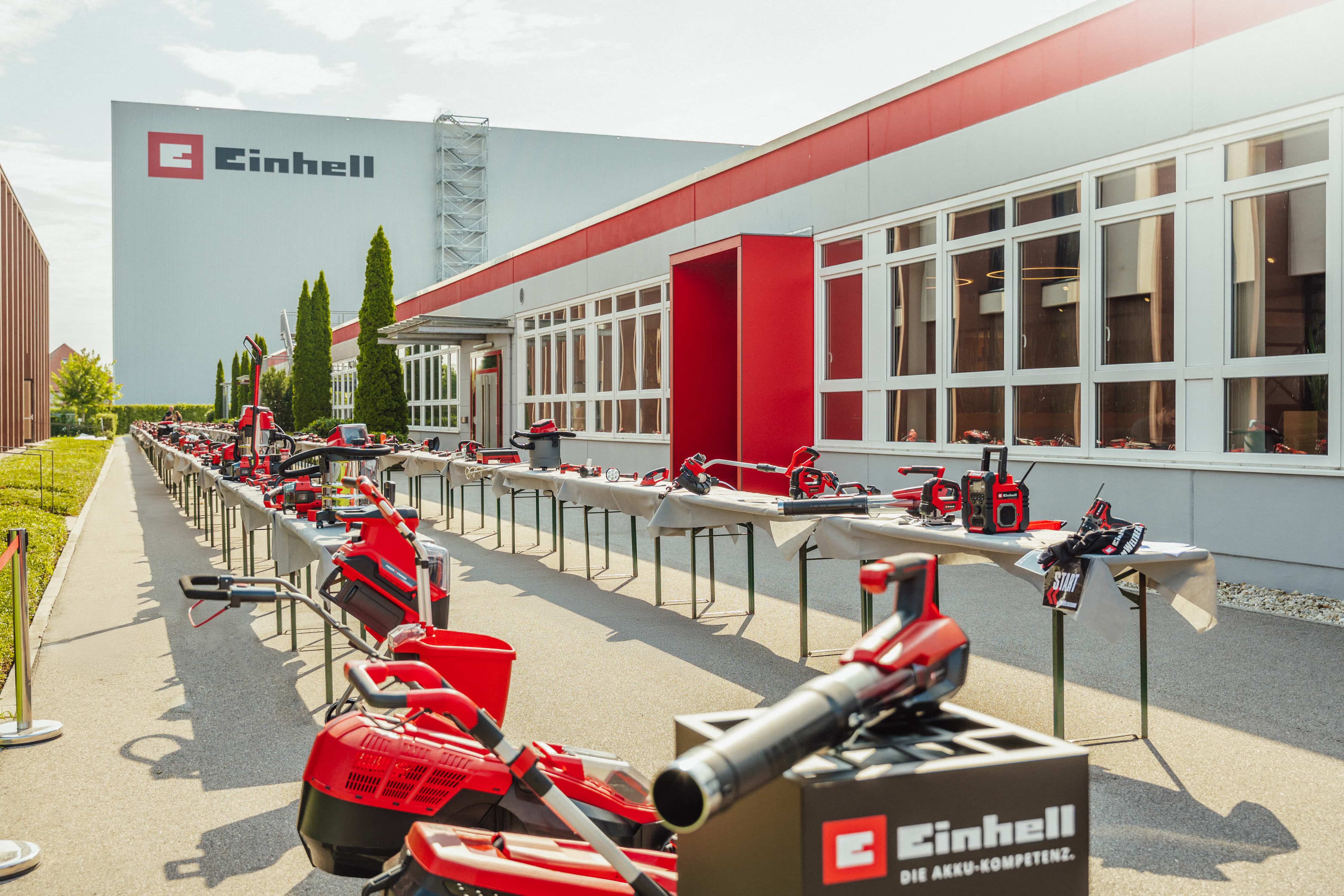 Long row of tables with various Einhell cordless tools on the outdoor area between company buildings.