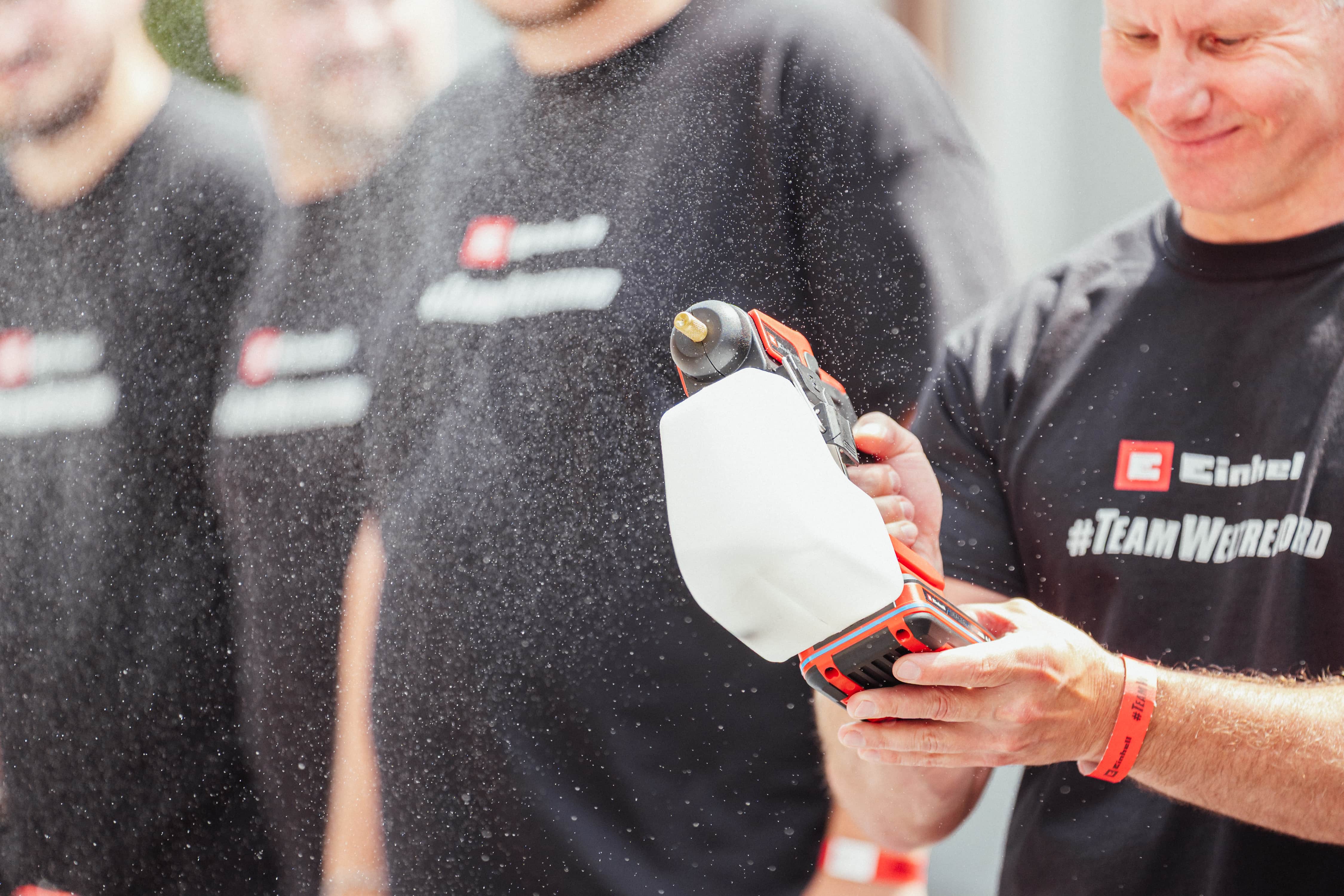 A man in a "#TeamWeltrekord" shirt holds an Einhell cordless spray system emitting fine mist.