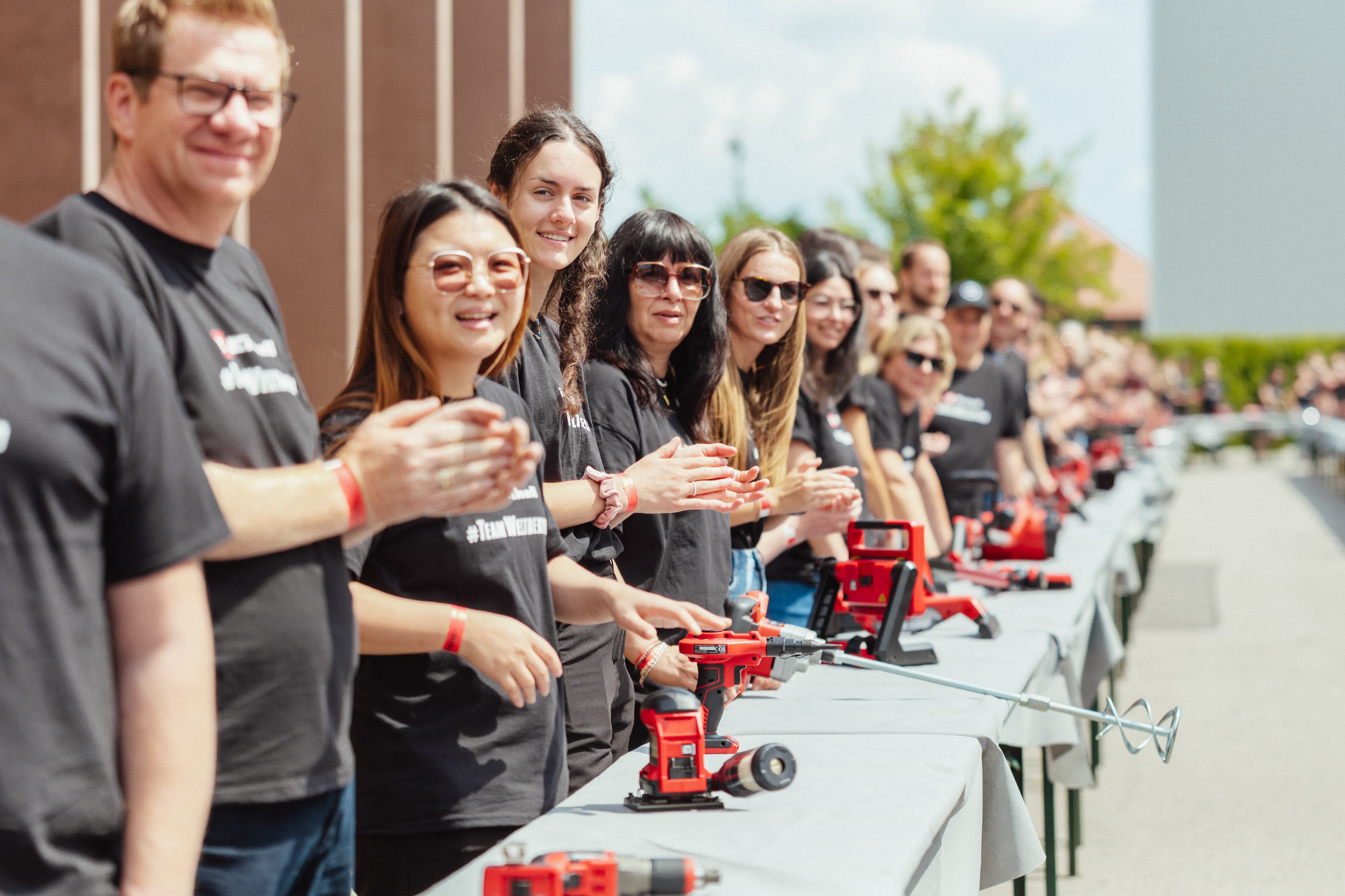 Einhell employees applaud beside a long row of cordless tools outdoors.