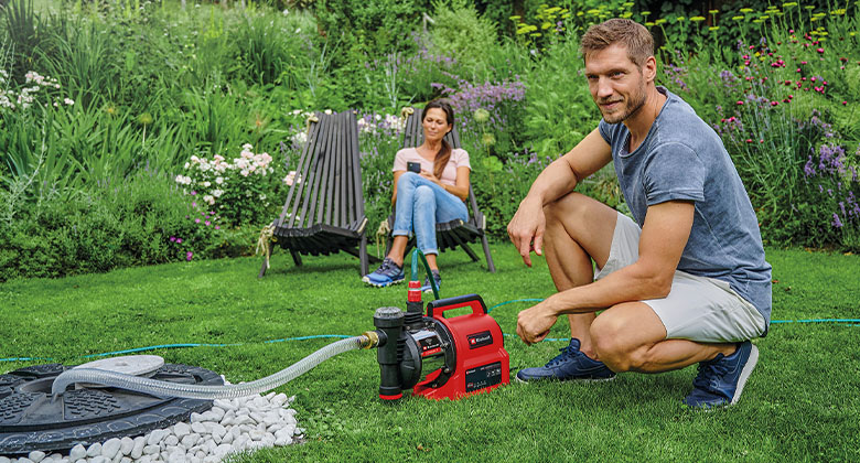 A man kneeling in the garden next to a garden pump connected to an underground water tank. In the background, a woman is sitting in a garden chair.