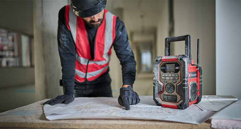 Bauarbeiter mit Helm und Warnweste zeigt auf einen Bauplan, neben ihm steht ein rotes Baustellenradio.