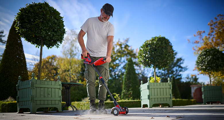Ein Mann reinigt mit dem Einhell Akku-Fugenreiniger Pflasterfugen auf einer Terrasse im Garten.
