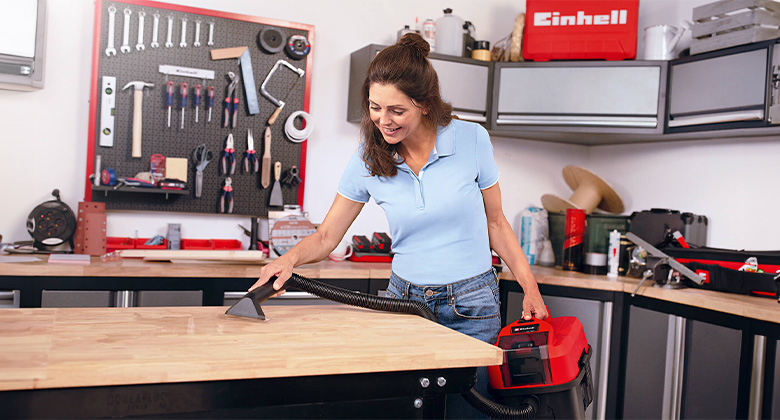 A woman uses an Einhell cordless wet/dry vacuum to suction water from a workbench in a workshop