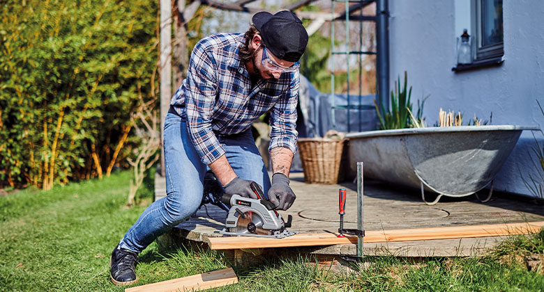 Man kneeling on his terrace and cutting a wooden slat with a hand-held circular saw